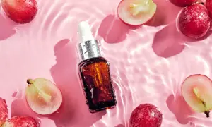 Odkryj naturalne olej z pestek winogron właściwości. Serum w buteleczce z pipetą otoczone świeżymi winogronami na różowym tle z falami wody.