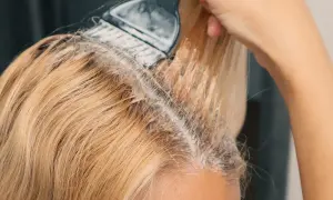 Ręka z pędzlem nakłada produkt do rozjaśniania włosów w domu. Widać blond pasma i początek zabiegu.