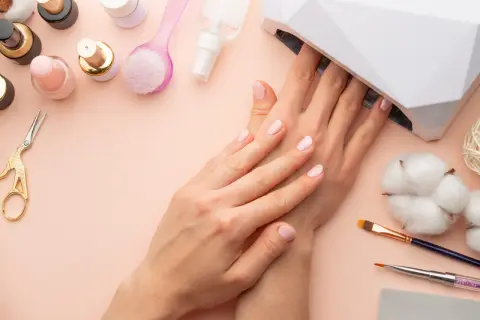 Delikatne paznokcie hybrydowe w ciąży, utwardzane w lampie UV. Wokół akcesoria do manicure: lakiery, pędzelki, nożyczki.