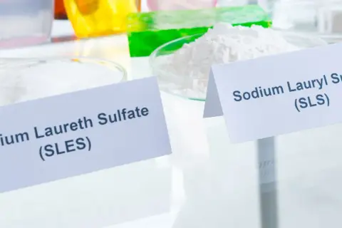 Białe proszki w naczyniach laboratoryjnych, jeden oznaczony jako Sodium Laureth Sulfate (SLES), drugi jako Sodium Lauryl Sulfate (SLS).
