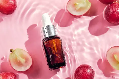 Odkryj naturalne olej z pestek winogron właściwości. Serum w buteleczce z pipetą otoczone świeżymi winogronami na różowym tle z falami wody.
