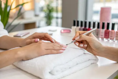 Dłonie podczas zabiegu manicure. Stylistka pielęgnuje paznokcie klientki, przygotowując je do malowania. W tle rząd lakierów.