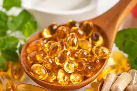 Łyżka pełna kapsułek z płynnym złotem – to suplementacja omega 3. Obok zielone liście i inne suplementy.