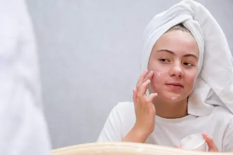 Dziewczyna z ręcznikiem na głowie nakłada krem na twarz. Witamina A pod oczy może pomóc w pielęgnacji skóry.
