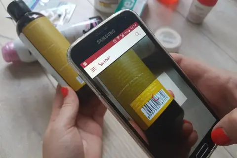 Ręce trzymają smartfon z otwartą aplikacją do sprawdzania składu kosmetyków. Na ekranie widoczny jest skaner kodu kreskowego skierowany na etykietę butelki.