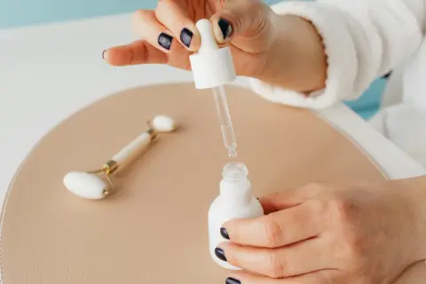 Dłoń z ciemnym lakierem do paznokci aplikuje serum z kwasem salicylowym i niacynamidem. Obok leży jadeitowy roller.