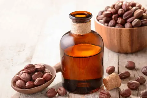 Bursztynowa buteleczka z olejkiem jojoba na twarz, obok drewniana łyżka z orzechami jojoba i miseczka pełna nasion.