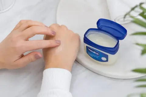 Dłoń rozsmarowuje wazelinę, sprawdzając, czy wazelina nawilża. Słoiczek Vaseline stoi obok.