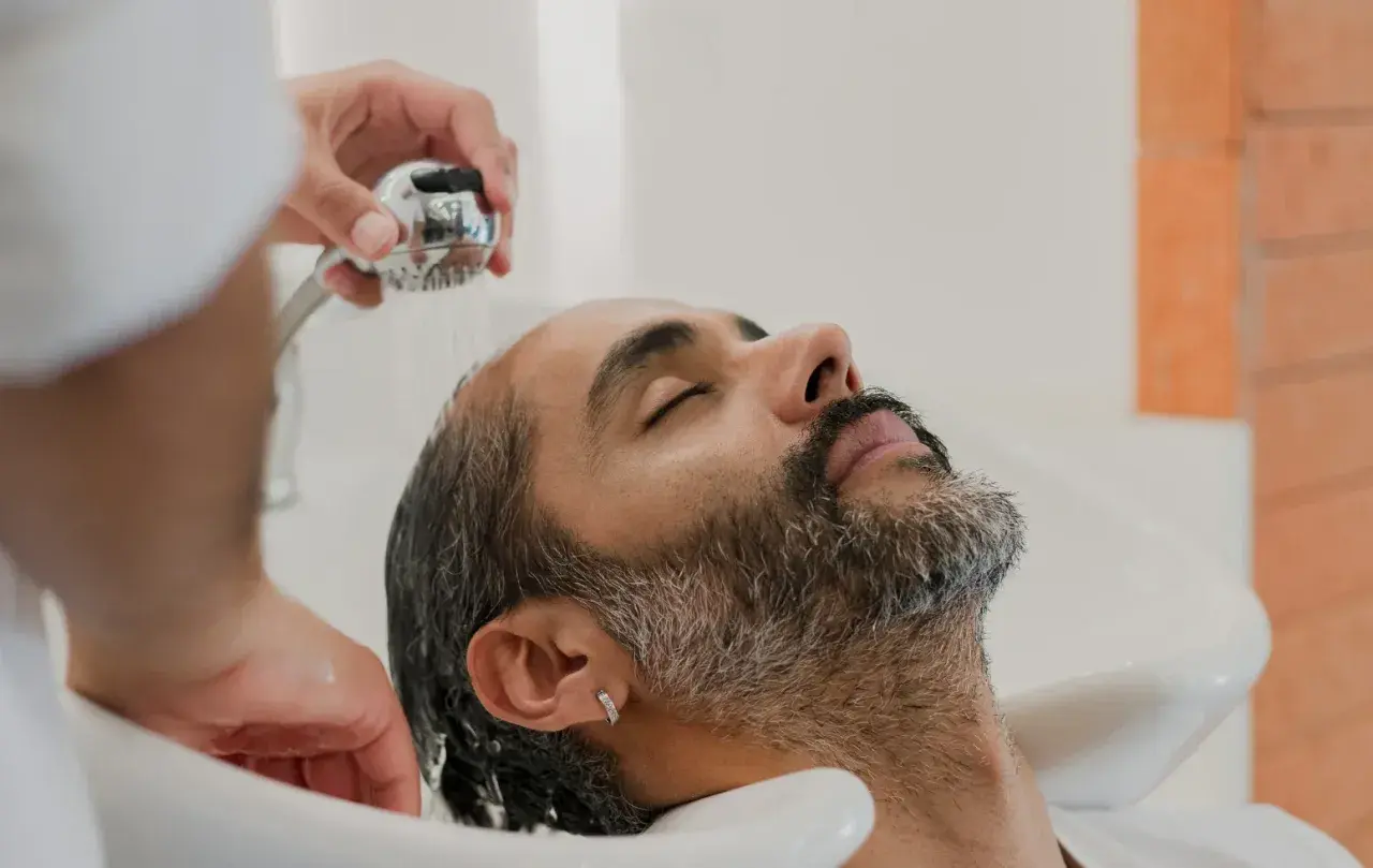 Mężczyzna z zarostem i kolczykiem w uchu myje włosy po przeszczepie. Delikatne strumienie wody spływają po jego twarzy.